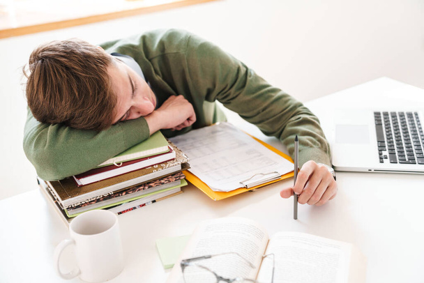 Зображення втомленого студента молодого хлопця за столом у приміщенні, який спить на книгах
. - Фото, зображення