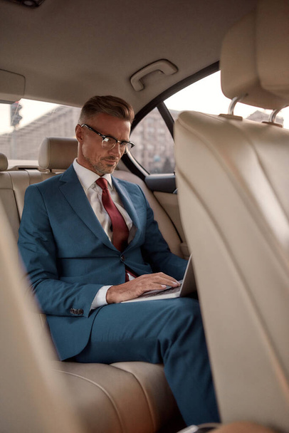Never stop working. Serious mature businessman in full suit working on his laptop while sitting in the car - Valokuva, kuva