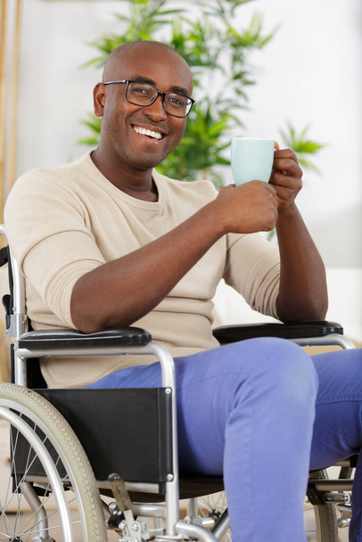 mature man in wheelchair drinking coffee near window indoors - Foto, imagen