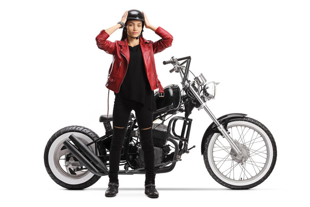 Full length portrait of a young female biker standing and putting on a helmet next to a chopper motorbike isolated on white background - Photo, Image