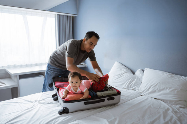 Dad put his clothes into a suitcase with a cute baby lying in an open suitcase - Foto, Bild
