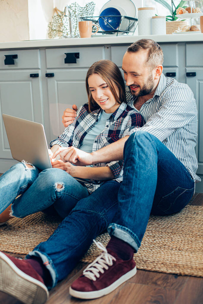 Beautiful couple of two young people sitting on the kitchen floor and pointing to a laptop screen - Photo, image
