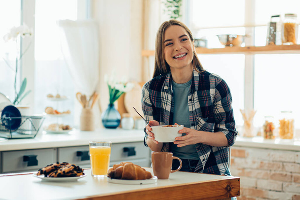 Mirthful young woman holding a bowl and smiling. Breakfast on the table in front of her - Photo, image
