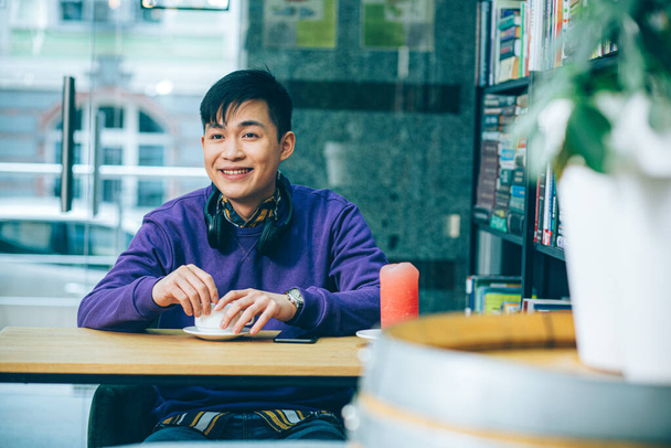 Cheerful young man sitting at the table with a cup of coffee and looking away with a smile - 写真・画像