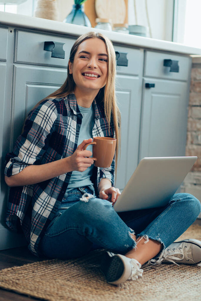 Smiling lady sitting on the floor with a laptop and holding a mug while looking away - Foto, Bild