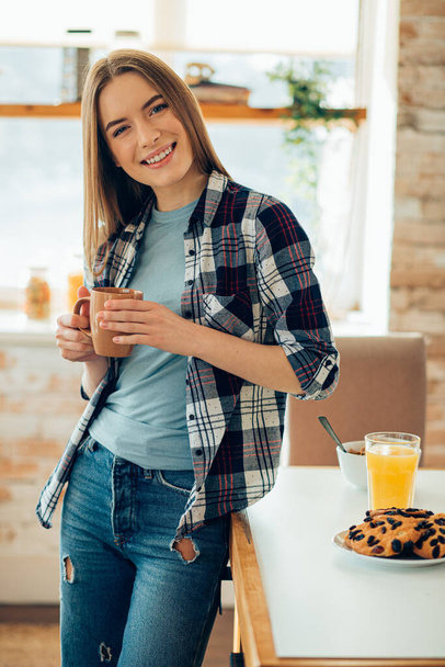 Usmívající se žena v neformálních šatech, stojící u stolu a držící hrnek - Fotografie, Obrázek