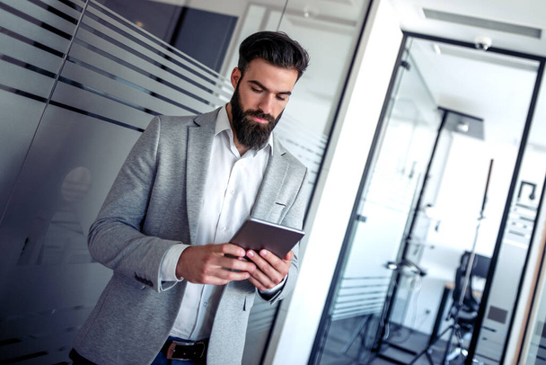 Jonge zakenman werkt online met een digitale tablet terwijl hij in kantoor staat. - Foto, afbeelding