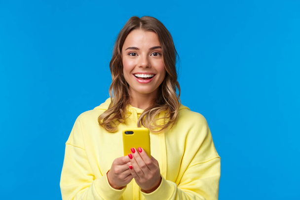 Close-up portrait excited smiling cute european female with blond haircut, holding mobile phone and looking camera entertained, standing blue background messaging, talking with friends online - Fotografie, Obrázek