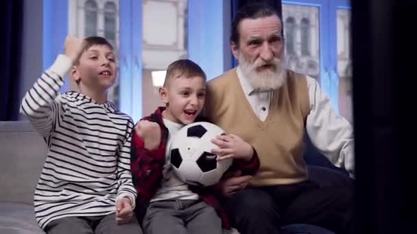 Front view of attractive exciting bearded senior grandfather with his teenage grandsons which watching sport game on tv and shouting - Materiał filmowy, wideo