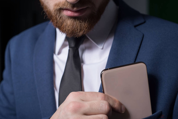 Close-up, studio dramatic portrait of a handsome bearded man, in a business suit, holding a new smartphone in his hand, against a tuxedo background. On a gray background. Office Style. Business design and style. New fashionable smartphone - Photo, Image