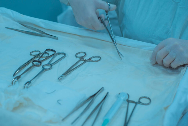 Κοντινό πλάνο, τα χέρια ενός χεριού νοσοκόμων, ένας βοηθός χειρουργού παίρνει ένα χειρουργικό όργανο από ένα αποστειρωμένο τραπέζι. Σε ένα αποστειρωμένο χειρουργείο. Αφαίρεση ενός κακοήθους όγκου του αυτιού με νυστέρι, συρραφή μαλακών ιστών, ηλεκτροπήξη, διακοπή αιμορραγίας - Φωτογραφία, εικόνα