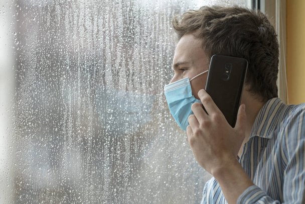 Junger Mann mit Gesichtsmaske, krank mit COVID-19, unter Quarantäne in einem Krankenhaus, beunruhigt am Telefon.  - Foto, Bild