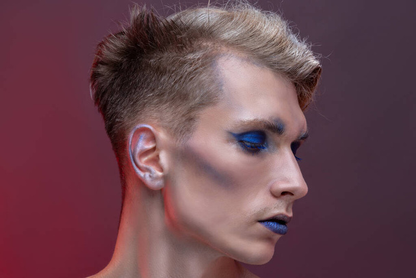 Close-up beauty portrait of a young man with blond hair on a red background. Professional makeup, lips and eyes with blue eyeshadows and lipstick, concept of face and hair care and natural clean skin - Fotoğraf, Görsel