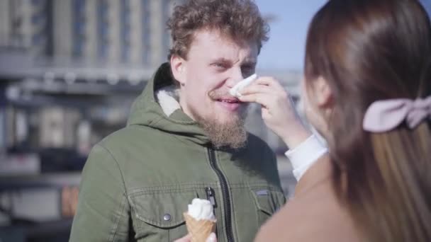 Unrecognizable brunette woman cleaning boyfriends nose dirty with ice cream. Positive young man smiling and looking at girlfriend with love. Shooting over shoulder. Lifestyle, dating, leisure. - Πλάνα, βίντεο