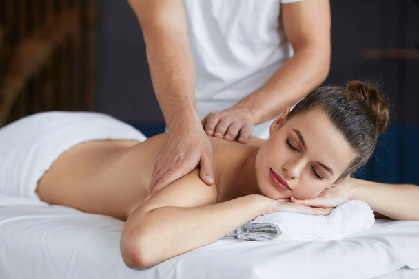 Jonge mooie vrouw genieten van rug en shouders massage in spa.Professionele massage therapeut is de behandeling van een vrouwelijke patiënt in appartement.Ontspanning, schoonheid, lichaam en gezicht behandeling concept.Home massage - Foto, afbeelding