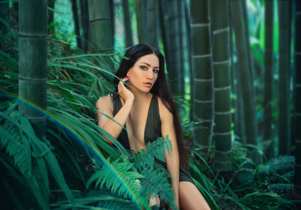 Forest fairy long black hair brunette enjoy green nature sexy emerald dress hand near face. Backdrop bamboo forest fern grass grove botanical garden tropics summer spring. Georgia fantasy art vacation - Fotó, kép