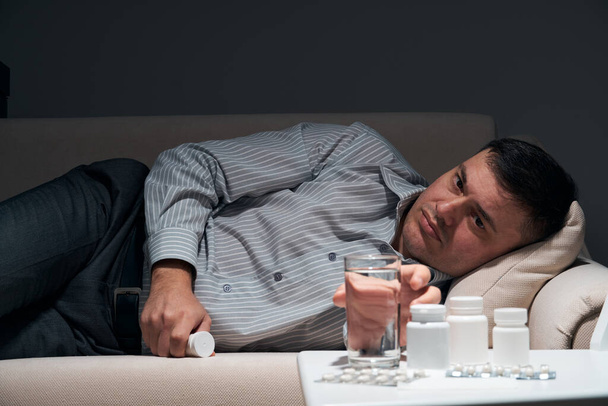 Empresário sobrecarregado está deitado no sofá e descansando. Comprimidos e copo de água perto dele
. - Foto, Imagem