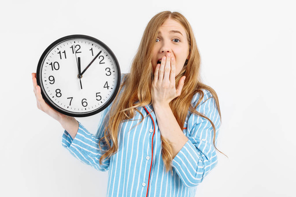 Portrait of shocked beautiful girl dressed in pajamas showing alarm clock isolated on white background - Foto, Imagem