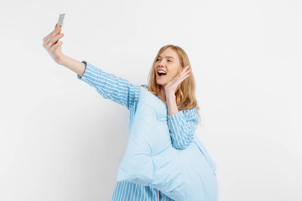 Excited beautiful young woman in pajamas holds a soft pillow and smiles while taking a selfie on a white background - Foto, immagini