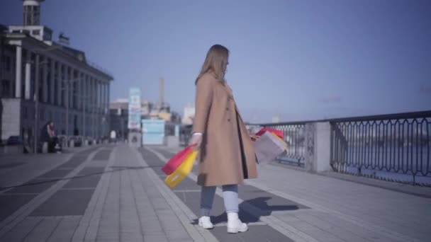 Wide shot of satisfied Caucasian woman in sunglasses and autumn coat spinning on river embankment with shopping bags and smiling at camera. Happy female shopaholic resting outdoors with purchases. - Záběry, video