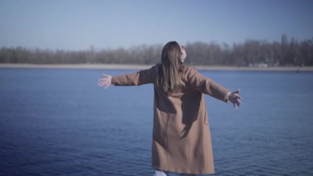 Back view of young Caucasian woman in autumn coat standing with hands stretched out. Positive brunette girl enjoying sunny day outdoors on river bank. Lifestyle, happiness, leisure. - Filmati, video
