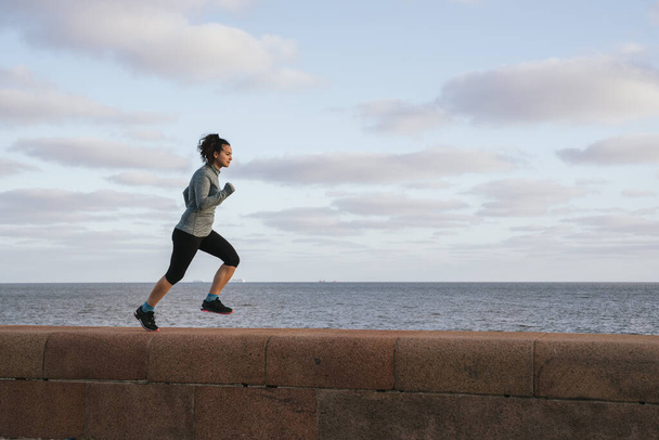 dziewczyna biegnie bokiem w prawo, w pobliżu morza, ćwicząc swoje ciało - Zdjęcie, obraz