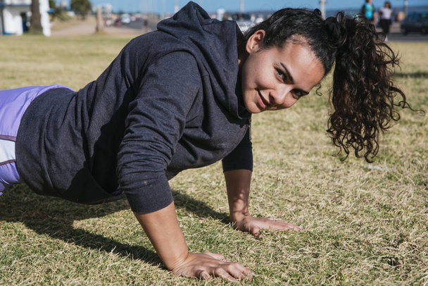 girl working out on the grass doing push ups, with her hair tied and looking at the camera - Photo, Image