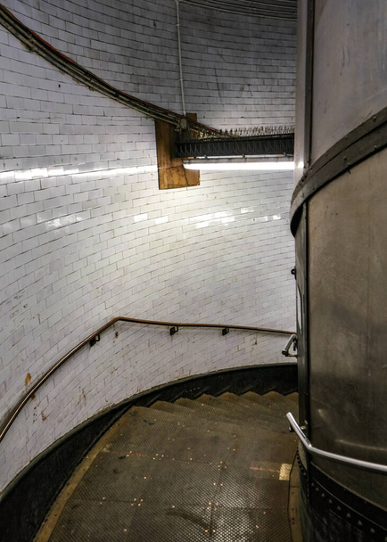 ネオンライトに照らされた暗く汚れた金属製の螺旋階段は、ロンドンのテムズ川の下のグリニッジ・フットトンネルにつながります。 - 写真・画像