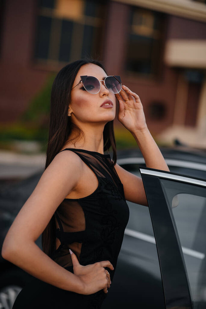 Stylish young girl stands near the car in a black dress. Business fashion and style - Фото, зображення