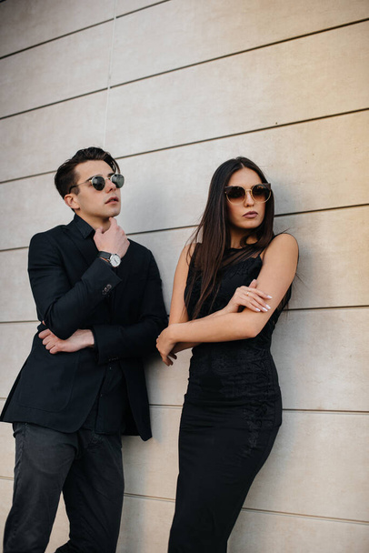 Un hermoso y elegante par de jóvenes vestidos de negro y gafas se colocan sobre el fondo de un edificio de oficinas al atardecer. Moda y estilo - Foto, Imagen
