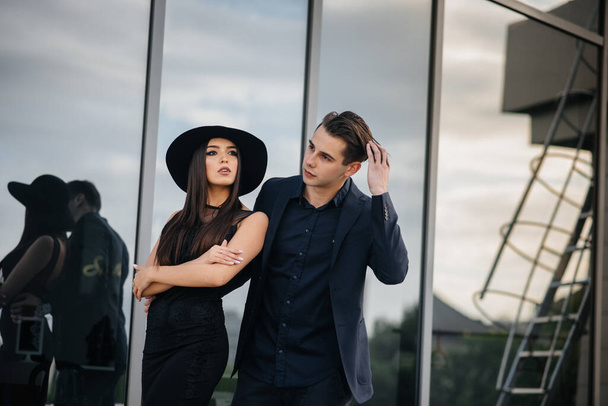 A beautiful, stylish pair of young people in black clothes and glasses stand against the background of an office building in the sunset. Fashion and style - Zdjęcie, obraz