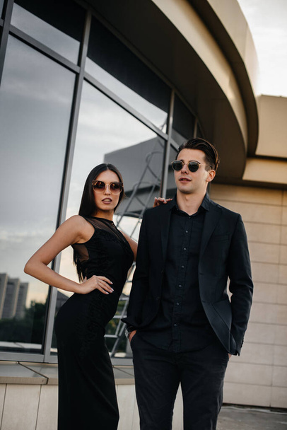 Um belo e elegante par de jovens com roupas e óculos pretos fica no fundo de um prédio de escritórios ao pôr-do-sol. Moda e estilo - Foto, Imagem