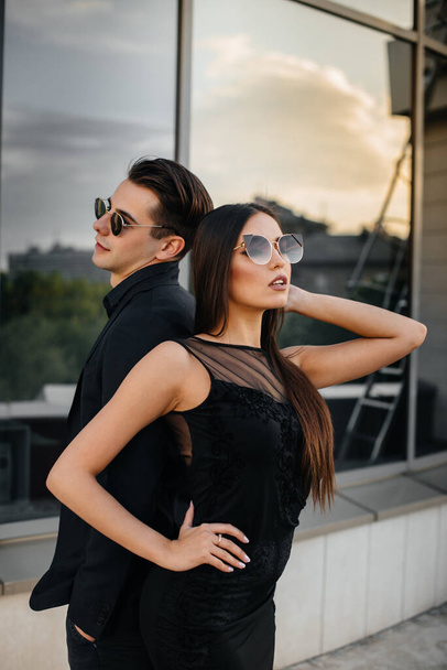 A beautiful, stylish pair of young people in black clothes and glasses stand against the background of an office building in the sunset. Fashion and style - Foto, Imagen
