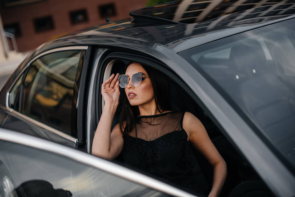 Stylish young girl sitting in a business class car in a black dress. Business fashion and style - Foto, afbeelding