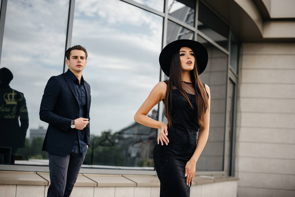 A beautiful, stylish pair of young people in black clothes and glasses stand against the background of an office building in the sunset. Fashion and style - Photo, Image