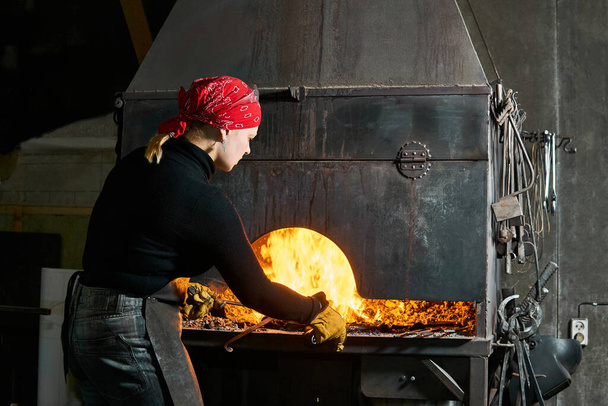 artista de metal feminino escolhe uma peça de um forno de ferreiro em chamas tradicional com pinça
 - Foto, Imagem