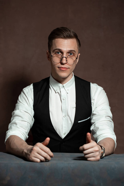 Beyaz gömlekli, siyah yelekli ve yuvarlak gözlüklü bir adam masada oturuyor. - Fotoğraf, Görsel