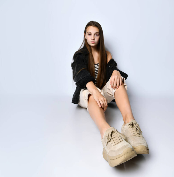 Teenage girl in striped t-shirt, black denim jacket, pants and sneakers. She is sitting on the floor isolated on white. Close up - Фото, изображение