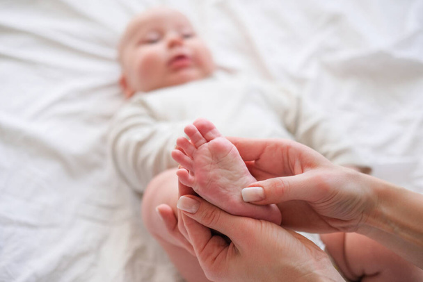 Baby feet in mother hands. Young caucasian woman makes massage for happy infant baby on white bed at home. Babycare, sport and happy motherhood. - Photo, Image