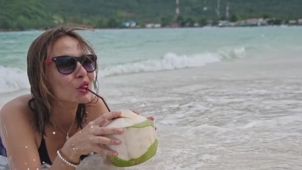 A young woman is lying on the beach in the sand.The girl is drinking coconut water. Coconut Cocktail. Smiling woman on a tropical island. Girl in the wave. - Footage, Video