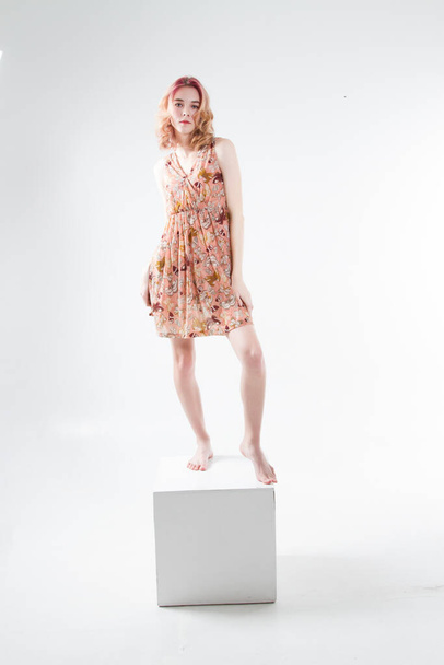 όμορφη νεαρή κοπέλα σε ένα σύντομο καλοκαιρινό φόρεμα στέκεται σε ένα λευκό κύβο - Φωτογραφία, εικόνα