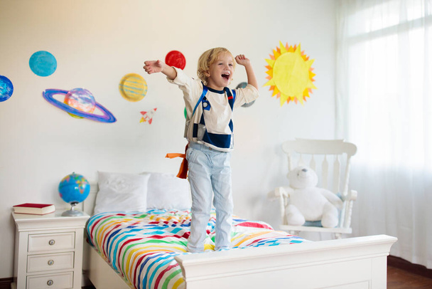 Дети играют в астронавтов. Мальчик в космическом костюме прыгает на кровати с ракетой. Солнечная система и декор планетной комнаты. Творческий ребенок, будущая профессия
. - Фото, изображение