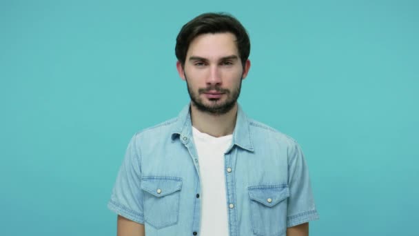 Cheerful happy young bearded guy in jeans shirt looking at camera with kind toothy smile, healthy content man expressing optimism, positive outlook. indoor studio shot isolated on blue background - Кадры, видео