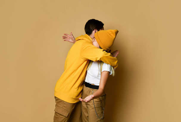 Young teens boy and girl in comfortable clothing, hats and sunglasses standing and embracing each other over yellow background - Fotoğraf, Görsel