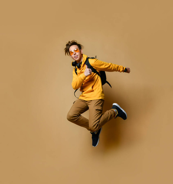 Young teen boy in comfortable clothing, sneakers, sunglasses and backpack jumping and feeling cool over yellow wall background - Φωτογραφία, εικόνα