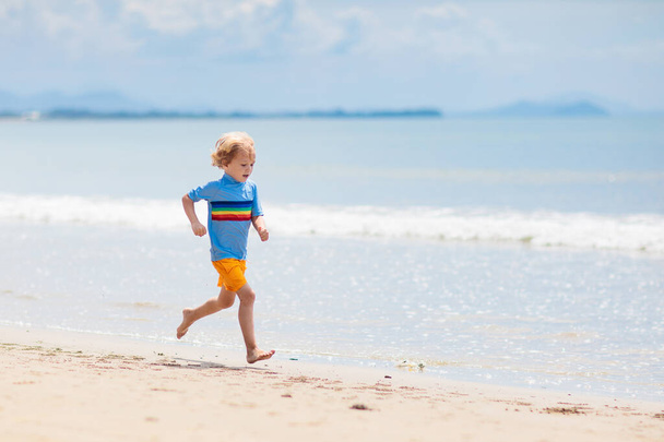 Παιδιά που παίζουν σε τροπική παραλία. Τα παιδιά κολυμπούν και παίζουν στη θάλασσα στις καλοκαιρινές οικογενειακές διακοπές. Η άμμος και το νερό διασκέδαση, προστασία από τον ήλιο για το μικρό παιδί. Μικρό αγόρι τρέχει και πηδάει στην ακτή του ωκεανού. - Φωτογραφία, εικόνα