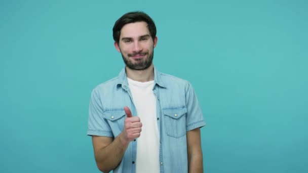 Joyful contented bearded guy in jeans shirt smiling pleased and showing thumbs up, male worker or employee satisfied with job, gesture of approval and success. studio shot isolated on blue background - Video, Çekim