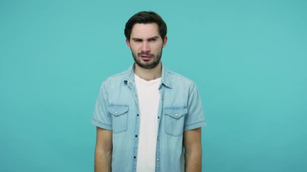Displeased bearded guy in jeans shirt pinching his nose holding breath, expressing disgust for unpleasant smell, bad breath, avoiding stinky odor. indoor studio shot isolated on blue background - Felvétel, videó
