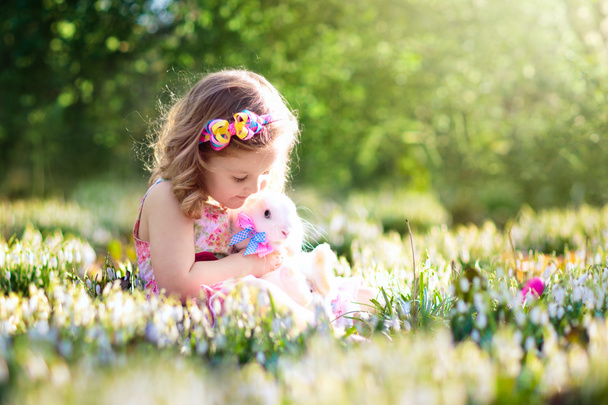 Roztomilá holčička, roztomilé kudrnaté batole v barevné letní šaty, hrál s skutečný králík, baví s její domácí bunny v krásné zahradě s první jarní květy sněženek - Fotografie, Obrázek