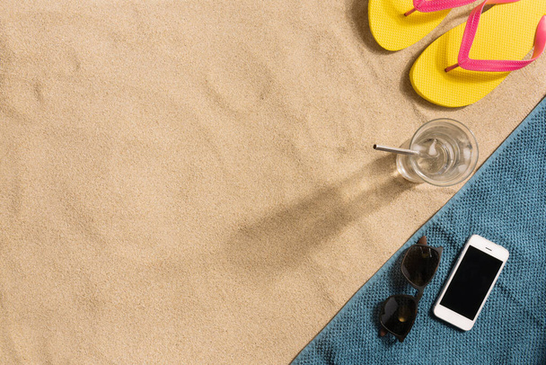 Summer vacation composition. Flip flops, smartphone and glass of water on sand background. Harsh light with shadows. Summer background. Border composition made of towel - Φωτογραφία, εικόνα